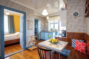 A cozy room with a bed, a dining table with fruit, a wooden chair, a kitchen area, and a window with curtains.