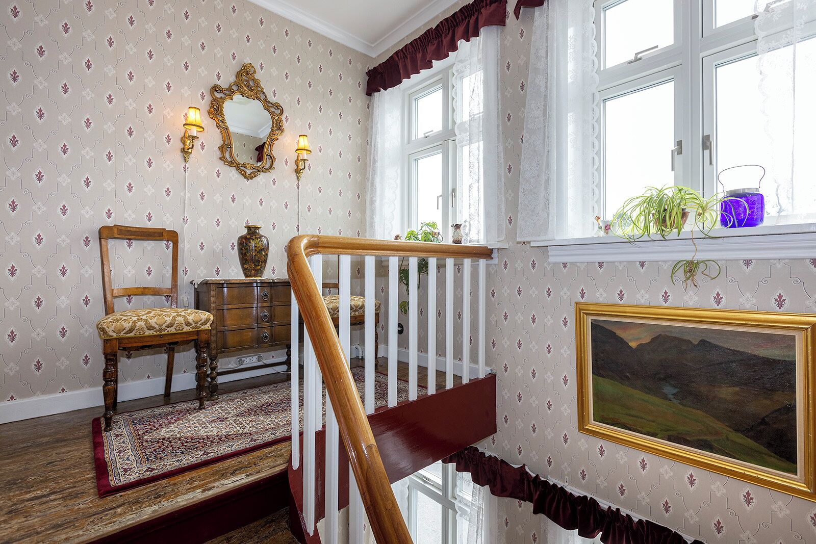 A room with patterned wallpaper, a wooden chair, a dresser, a mirror, a staircase with a railing, a window with plants, and a framed landscape painting.