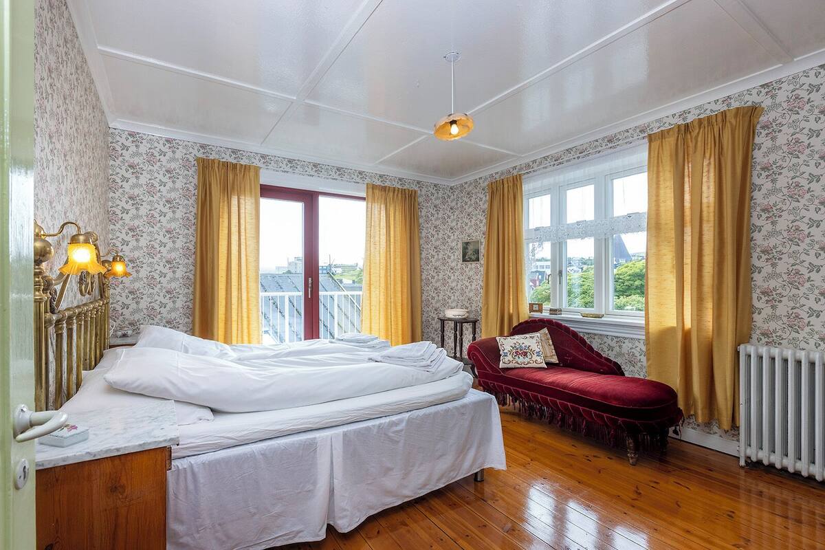 A bedroom with a bed, a red sofa, a window with curtains, and a radiator.