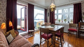 A room with a wooden dining table, red upholstered chairs, a patterned sofa, and a view of a building through large windows. - Downtown - Retro Decor - Stunning Views - Terrace (Tórshavn)