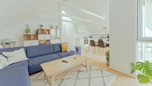 A modern living room with a blue sofa, a wooden coffee table, and a white rug. There is a kitchen area in the background with white cabinets and a dining table. - Stunning Views / Penthouse / Decorative / Parking (Nykobing Sjaelland)
