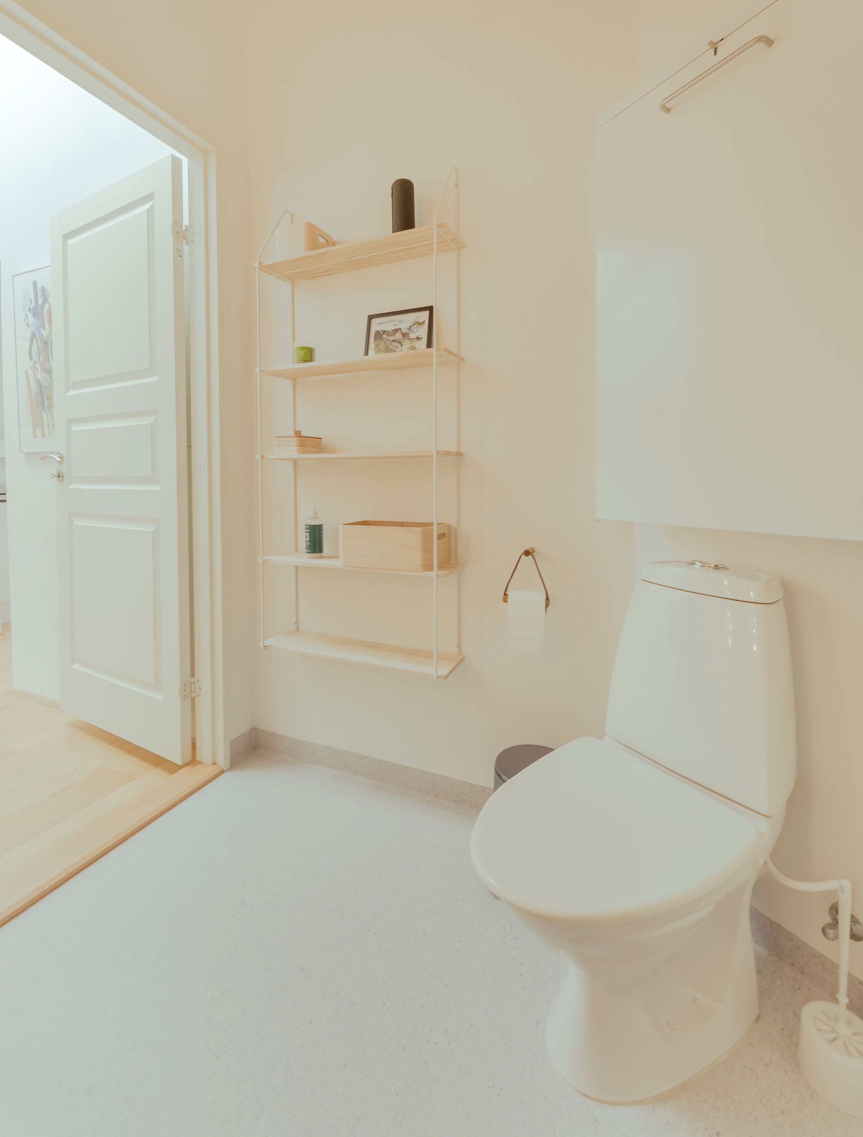 A bathroom with a white toilet, a shelf unit with various items, and a door leading to another room.