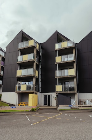Un edificio moderno de varios pisos con balcones, un pequeño espacio al aire libre para sentarse y un estacionamiento.