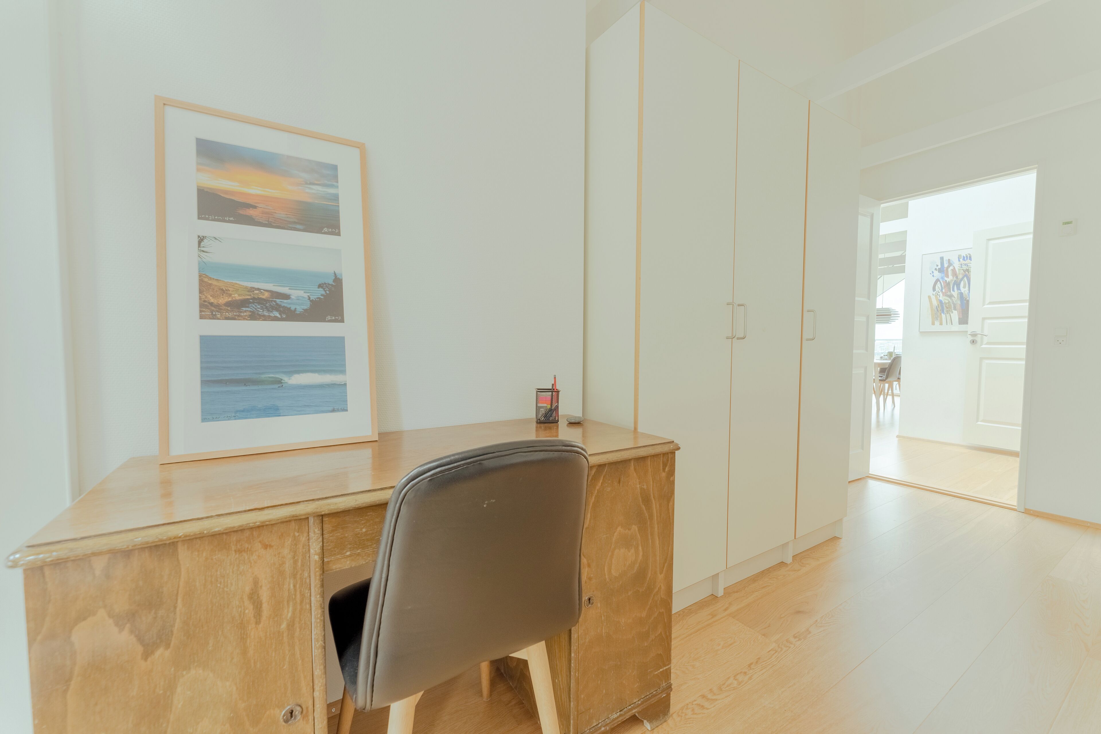 A modern room with a wooden desk, a framed picture of a coastal landscape, and a door leading to another room.