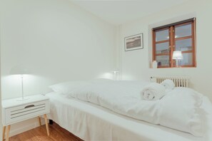 Cama bem arrumada com lençóis brancos e uma toalha enrolada, mesa de cabeceira branca com abajur, e janela com moldura de madeira e radiador branco abaixo.