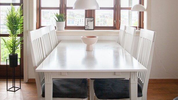 A dining area with a white table, white chairs, and two pendant lights. There is a window with a view of buildings and a potted plant on the left. - Aparthotel - Marina - Central - Tórshavn (Tórshavn)