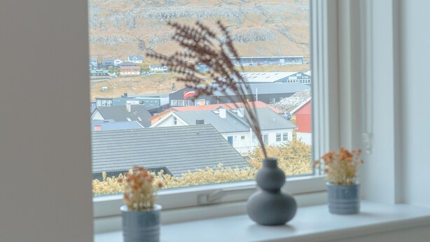 A window with a view of a town and hills, a gray vase with dried plants, and a gray couch.