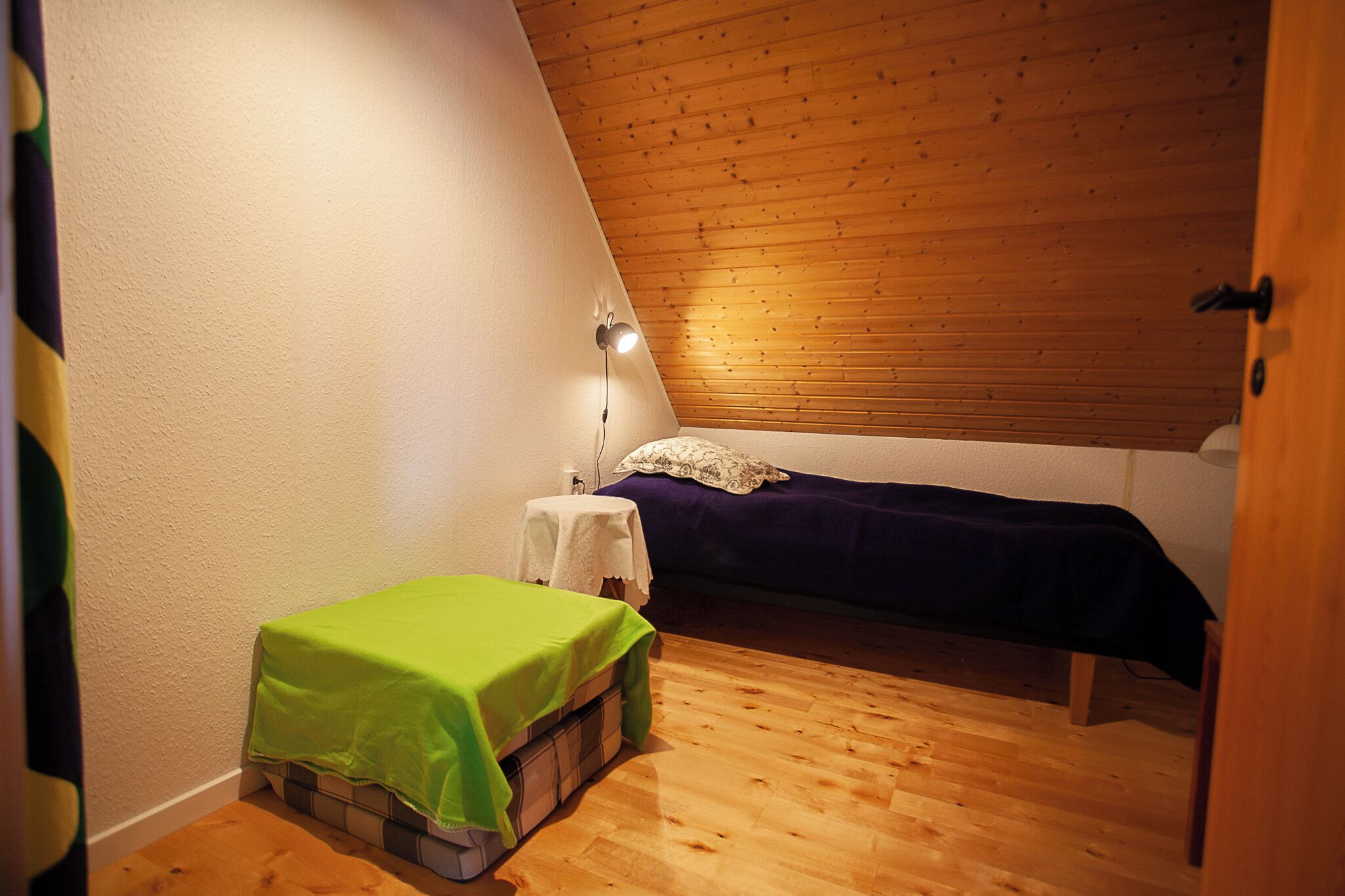 A small, cozy room with a sloped ceiling, a single bed with a green blanket, a bedside table with a lamp, and a wooden floor.