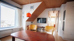 A dining area with a wooden table and chairs, a television, and a view of the city through a large window. - Tórshavn - Central - City & Ocean Views - 3Br (Tórshavn)