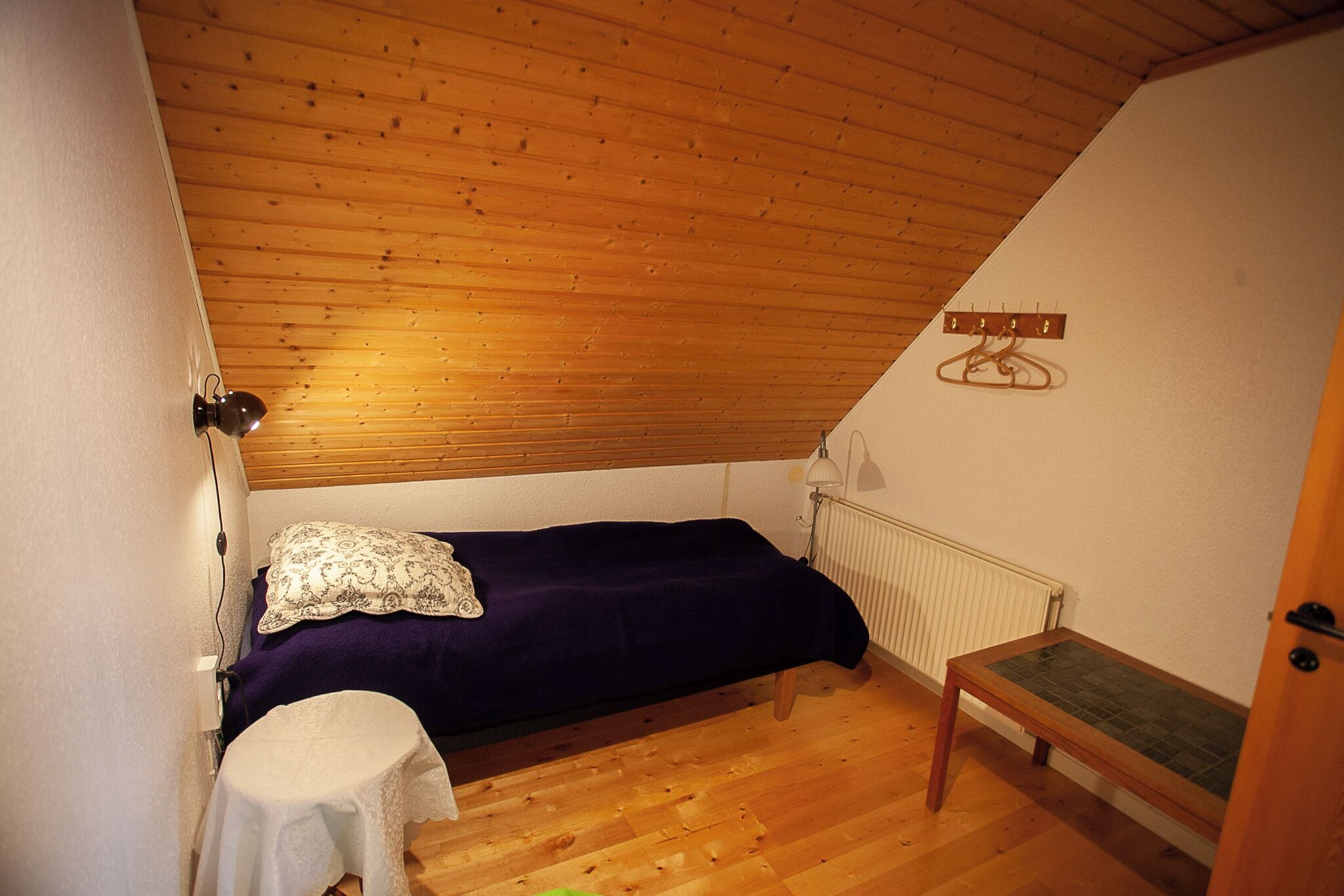 Um quarto pequeno e acolhedor com teto de madeira, uma cama simples com manta roxa, uma mesinha e um radiador.