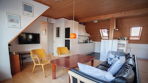 A cozy living room with a wooden ceiling, a sofa with cushions, a dining table with chairs, and a kitchen area with a sink and stove. - Tórshavn - Central - City & Ocean Views - 3Br (Tórshavn)