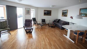 A spacious living room with a wooden floor, a grey sofa, a dining table with chairs, and a large window with a view of the outdoors. - Large Apartment With Fabulous View Of Tórshavn (Tórshavn)