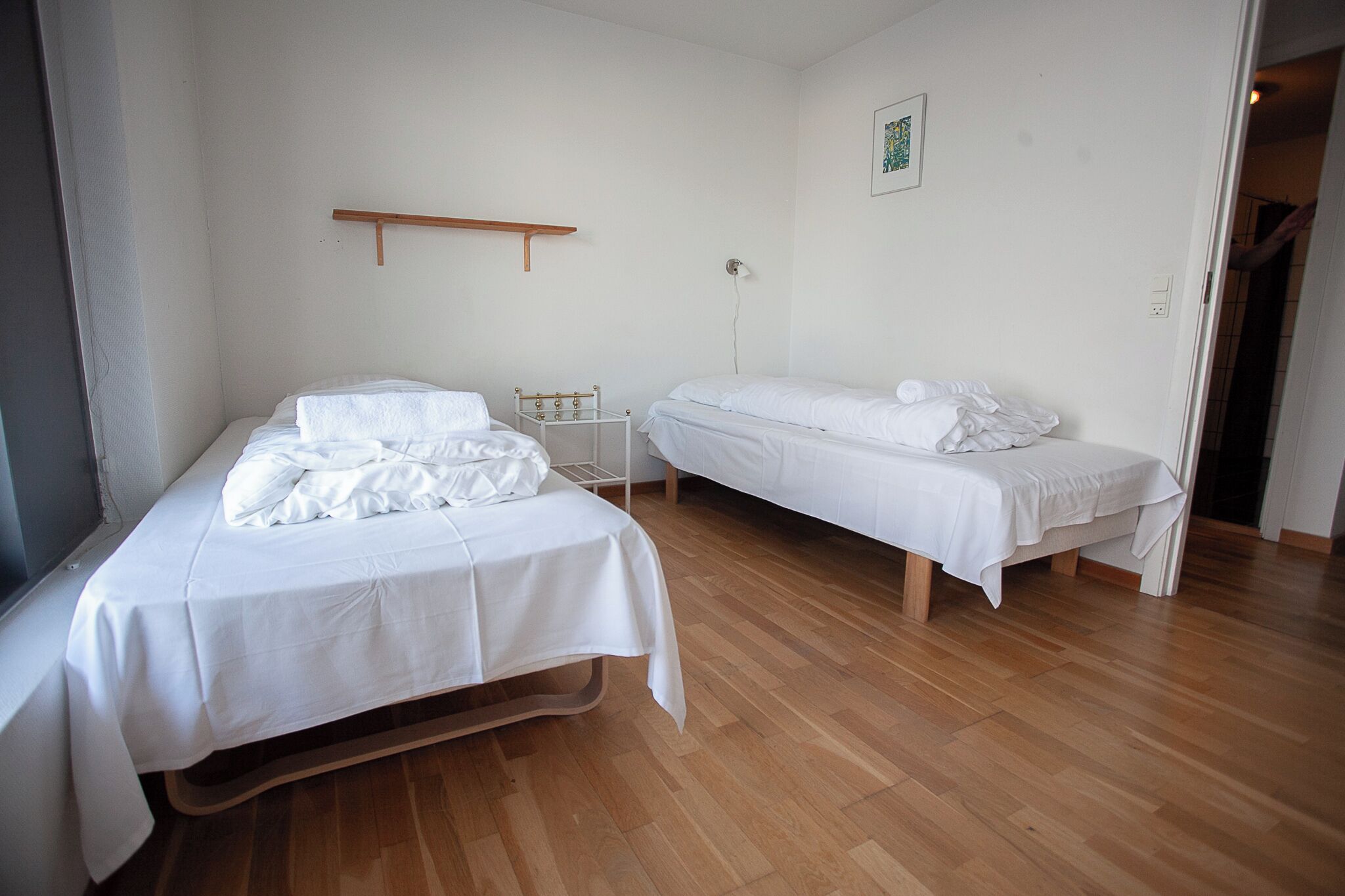 Una stanza con due letti, pavimento in legno, una mensola a muro e un unico punto luce.