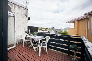 Um deck de madeira com mesa e cadeiras, um corrimão preto e vista para casas ao fundo.