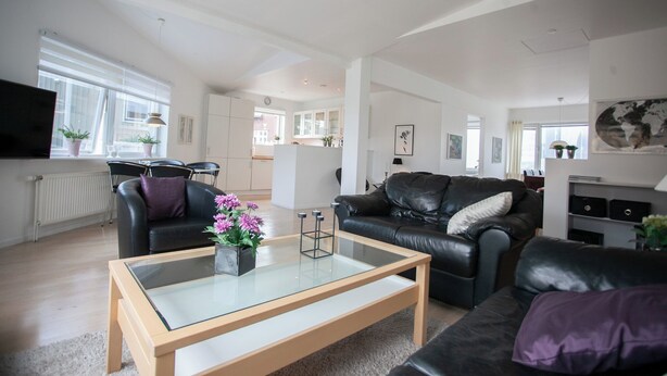 A modern living room with a glass coffee table, leather sofas, and a view of the kitchen and dining area.