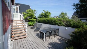 Eine Holzterrasse mit Tisch und Stühlen, eine kleine Treppe und Blick auf die umliegende Bepflanzung sowie Gebäude.