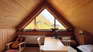 A room with a sloped ceiling, a wooden desk, a chair, a guitar, a radiator, and a window with a view of a hillside. - Idyllic Vacation Home With A Breathtaking View (Bøur)