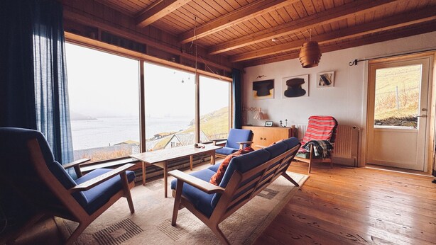 A room with wooden floors, a large window overlooking a body of water, a table, two chairs, and a cabinet with a picture on top. - Idyllic Vacation Home With A Breathtaking View (Bøur)