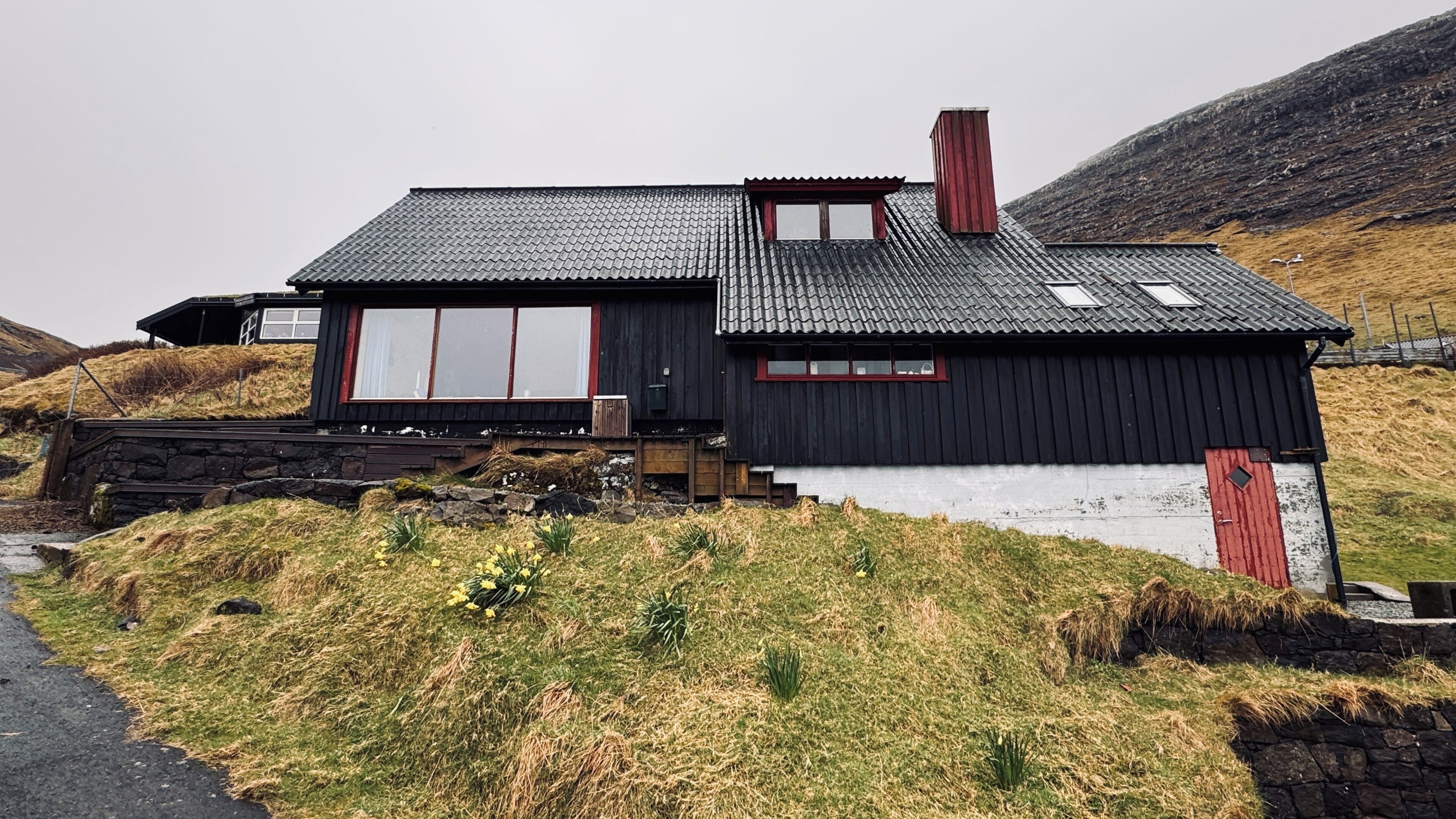 A black wooden house with a red door, a stone wall, and a grassy hill in the background.