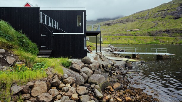 A black building with a staircase leading to a deck over water, surrounded by rocks and greenery.