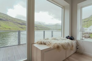 Ein Zimmer mit großen Fenstern, die auf einen See und Hügeln Blick bieten, eine Felldecke auf einer Bank und eine Lampe auf einem Fensterbrett.