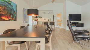 A modern kitchen with white cabinets, a dining area with a long table and chairs, and a TV on a stand.