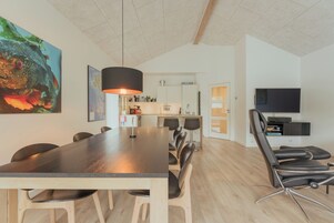 Eine moderne Küche mit weißen Schränken, ein Essbereich mit einem langen Tisch und Stühlen sowie ein Fernseher auf einem Schrank.