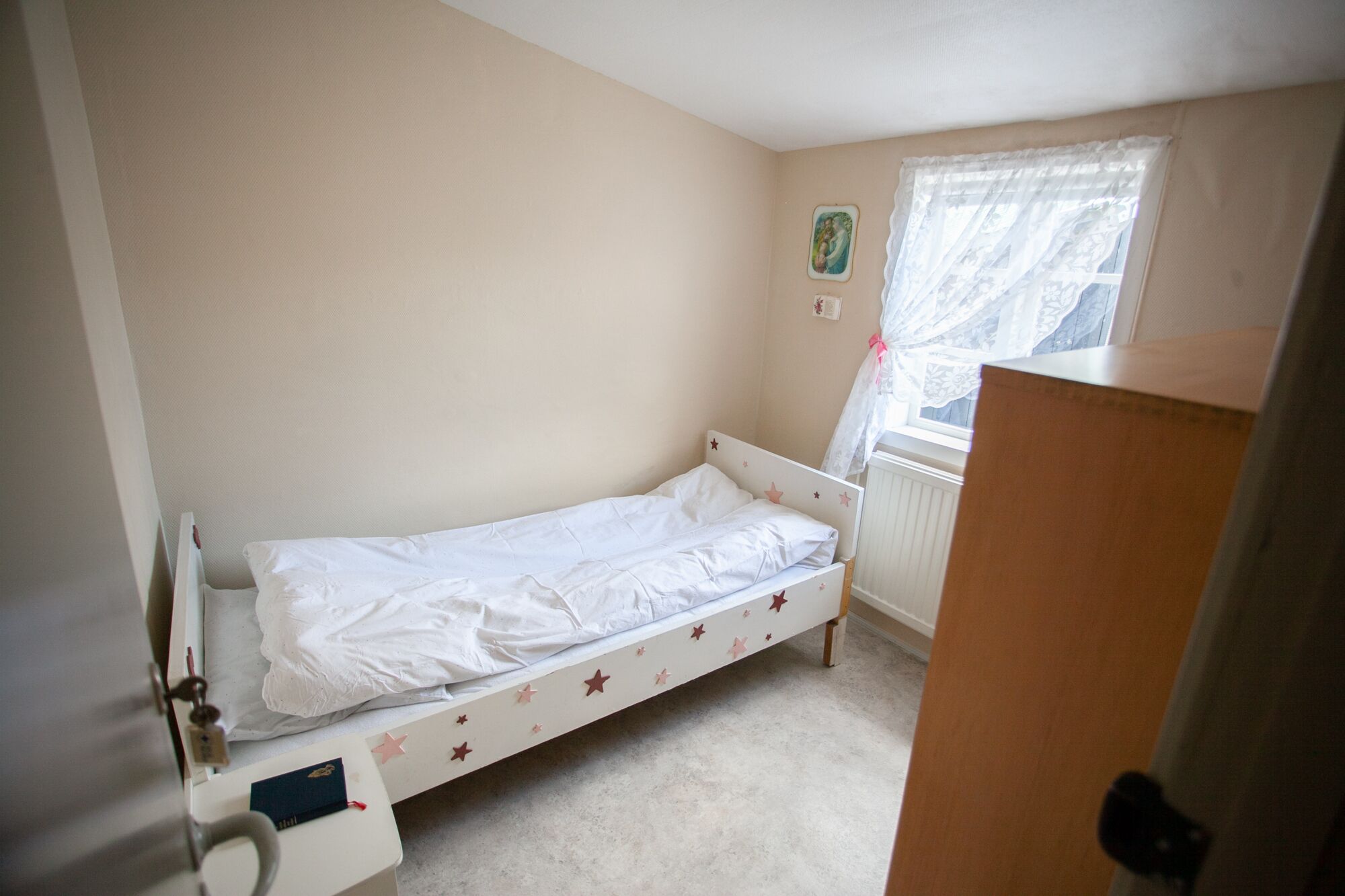 A small, single-bed room with a wooden headboard, a bedside table, and a window with a lace curtain.