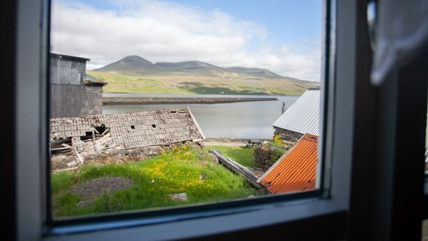 A view of a lake with mountains in the background, a building with a corrugated metal roof, and a stone wall. - Cozy House In The Idyllic Village Of Haldórsvík (Haldórsvík)