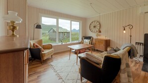 A cozy living room with wooden floors, a large window with a view of a house and mountains, a round wall clock, and a fireplace. - Authentic Cabin - Unique Location - In Nature - 2Br (Hellurnar)
