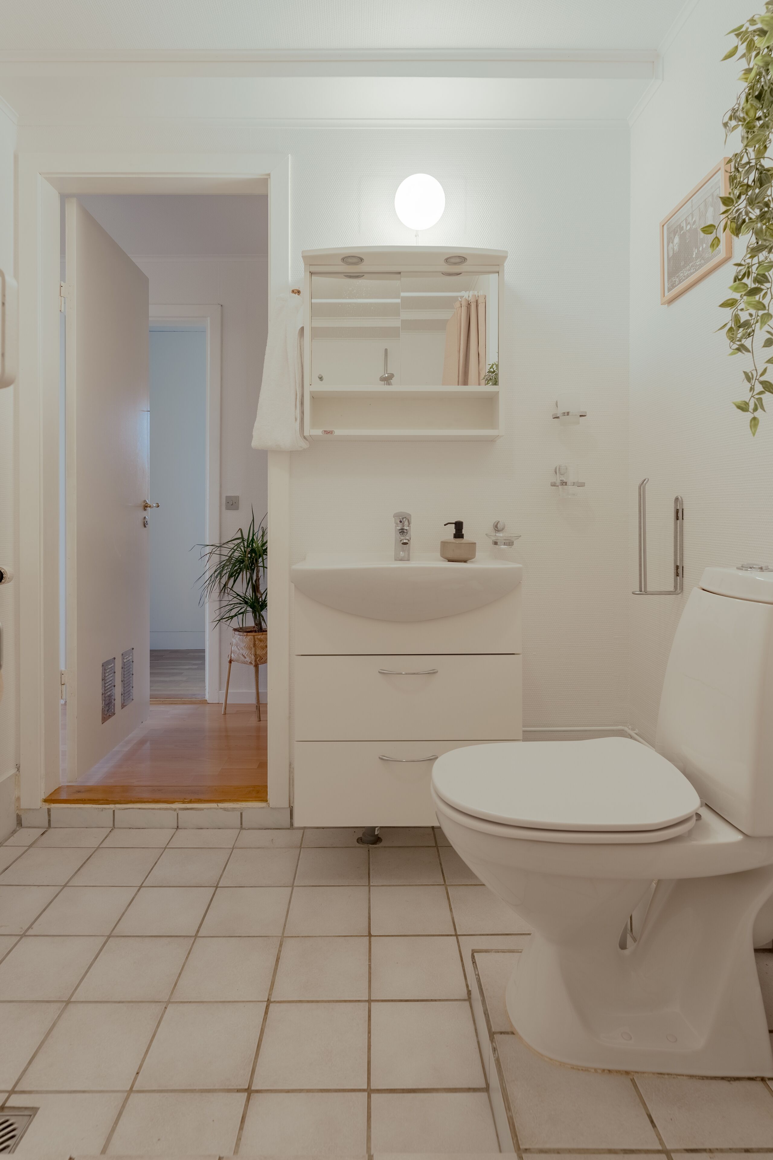 A compact bathroom with a white sink, toilet, and a small mirror. A potted plant is visible in the background.