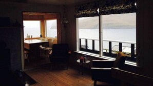 A cozy room with a wooden floor, a dining table with chairs, a small round table with a chair, and a large window overlooking a body of water. - Cottage With Stunning View Of The Sea And Islands. (Leynar)