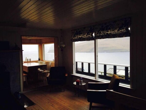 A cozy room with a wooden floor, a dining table with chairs, a small round table with a chair, and a large window overlooking a body of water.