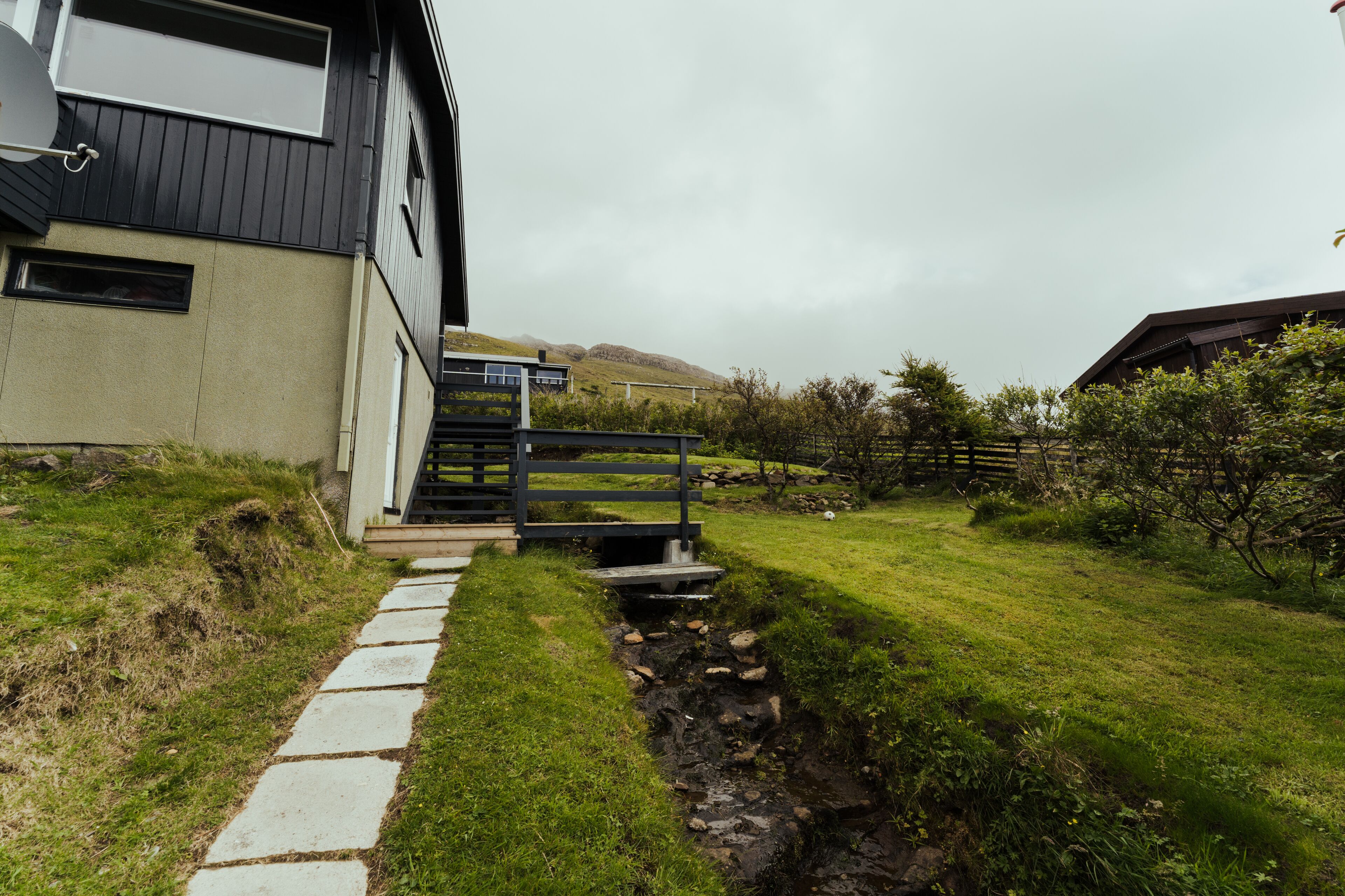 A pathway leading to a house with a small stream, surrounded by grass and trees.