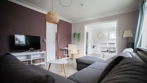 Uma sala moderna com um amplo sofá modular cinza, uma TV de tela plana sobre um suporte branco, uma mesa redonda pequena, cadeiras de vime e um abajur de vime.
