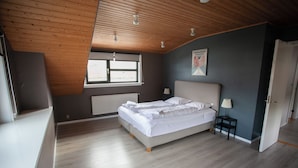 Quarto com teto de madeira, cama com roupa de cama branca, janela e radiador.