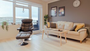 A modern living room with a leather chair, a sofa, and a coffee table. There is a large window with a view of the city, a clock on the wall, and two framed pictures. - Unique Boathouse - Central - Kajak - Fitness (Strendur)