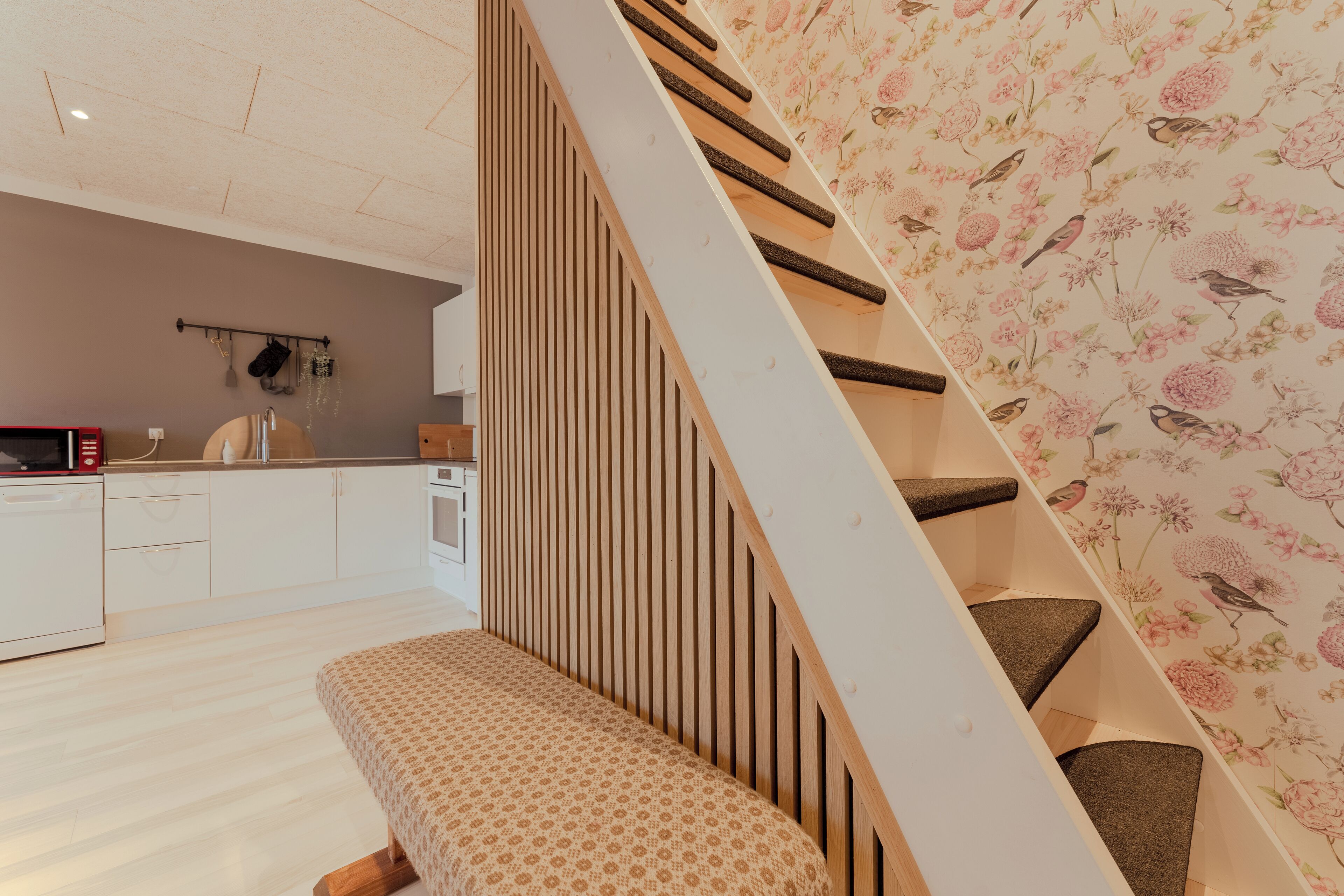 A modern kitchen with white cabinetry, a wooden bench, and a staircase with floral wallpaper.