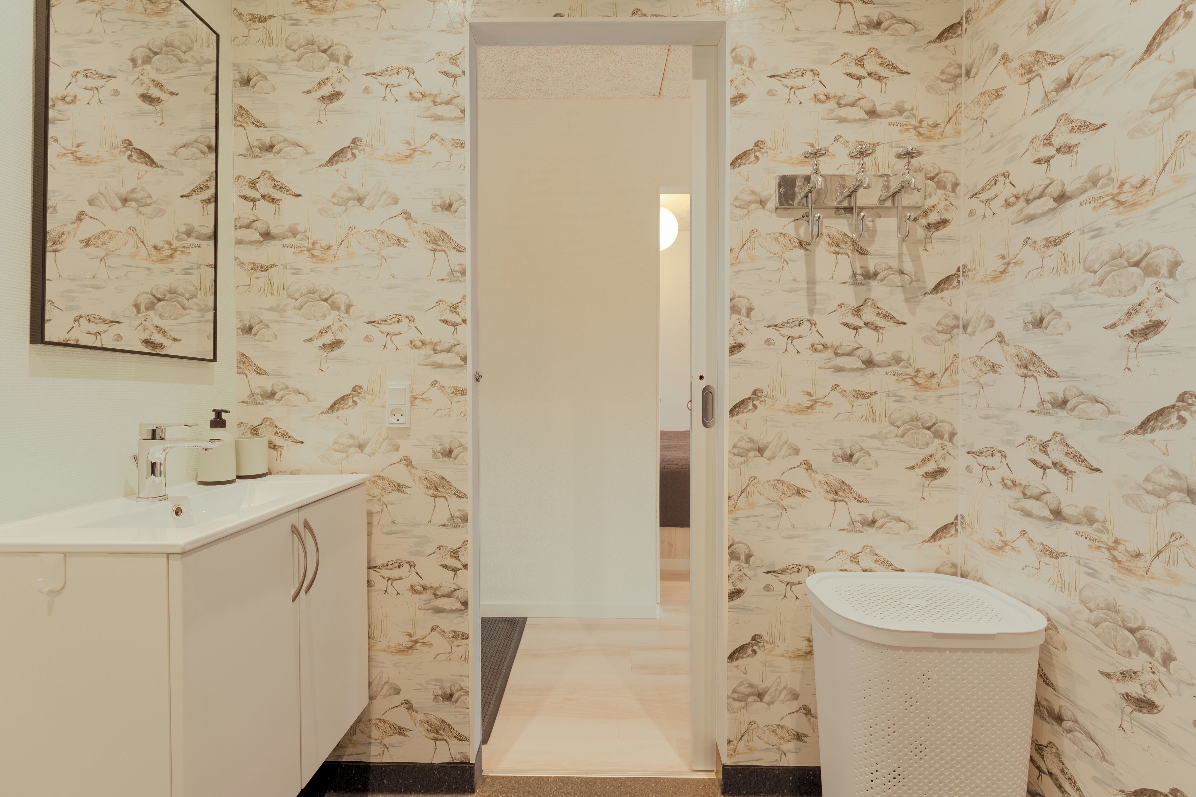 Une salle de bain avec un lavabo, un miroir et des toilettes. Les murs sont recouverts d’un papier peint à motifs d’oiseaux.