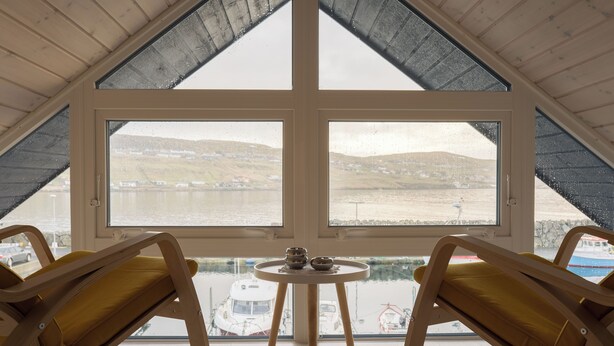 A room with a large window overlooking a body of water and hills. There are two wooden chairs with cushions and a small table with a bowl and a cup. - Unique Boathouse - Central - Kajak - Fitness (Strendur)