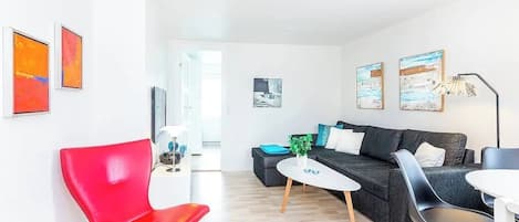 A modern living room with a red chair, a black sofa, and a white coffee table. There are abstract paintings on the walls and a lamp on a side table.
