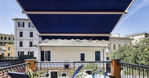Um terraço com mobiliário exterior, uma pérgula e vista para um edifício com varandas.