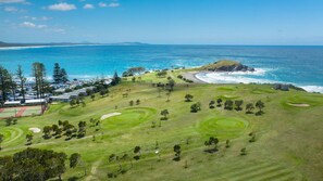 Un campo da golf con bunker, una distesa d'acqua e una catena montuosa in lontananza.