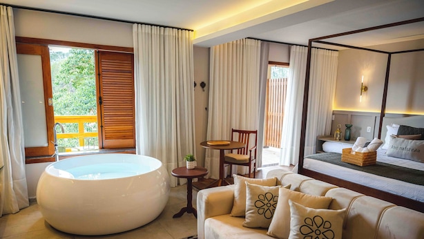 Une chambre d’hôtel moderne dotée d’une grande baignoire ronde, d’un canapé agrémenté de coussins décoratifs, d’une petite table ronde et d’un lit offrant une vue sur l’extérieur.