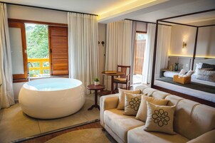 Una habitación de hotel moderna con una bañera redonda grande, un sofá con cojines decorativos, una mesita redonda y una cama con vistas al exterior.