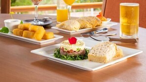 Une table dressée avec de la nourriture et des boissons, un verre de jus d’orange et un verre de bière, avec vue sur les arbres à l’extérieur.