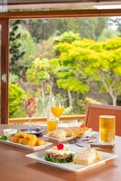Una mesa servida con comida y bebidas, un vaso de jugo de naranja y un vaso de cerveza, con vista a árboles afuera.