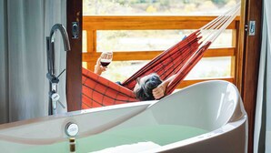 Une personne se détend dans un hamac près d’une grande baignoire, avec vue sur une terrasse en bois et la verdure environnante.