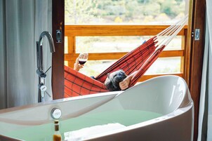 Una persona relajándose en una hamaca cerca de una bañera grande, con vista a una terraza de madera y vegetación.