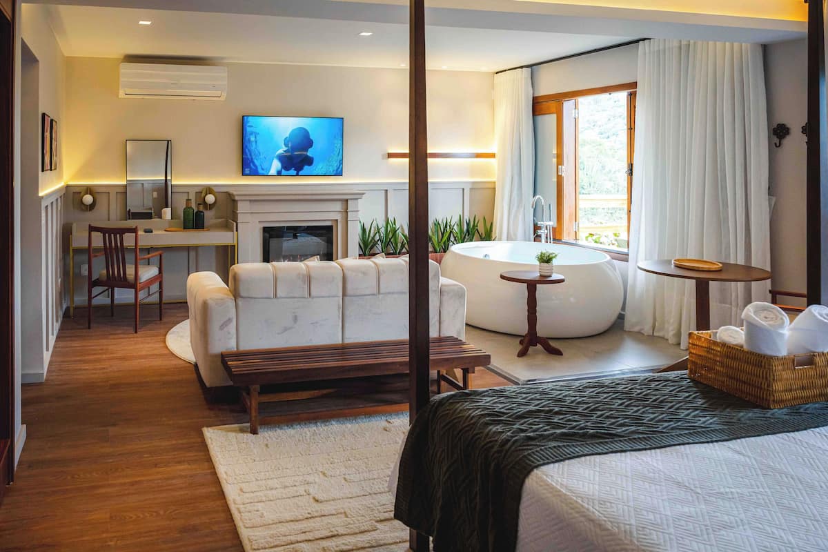 A modern hotel room with a sofa, a flat-screen TV, a fireplace, a large window with curtains, a round bathtub, and a small table with towels.
